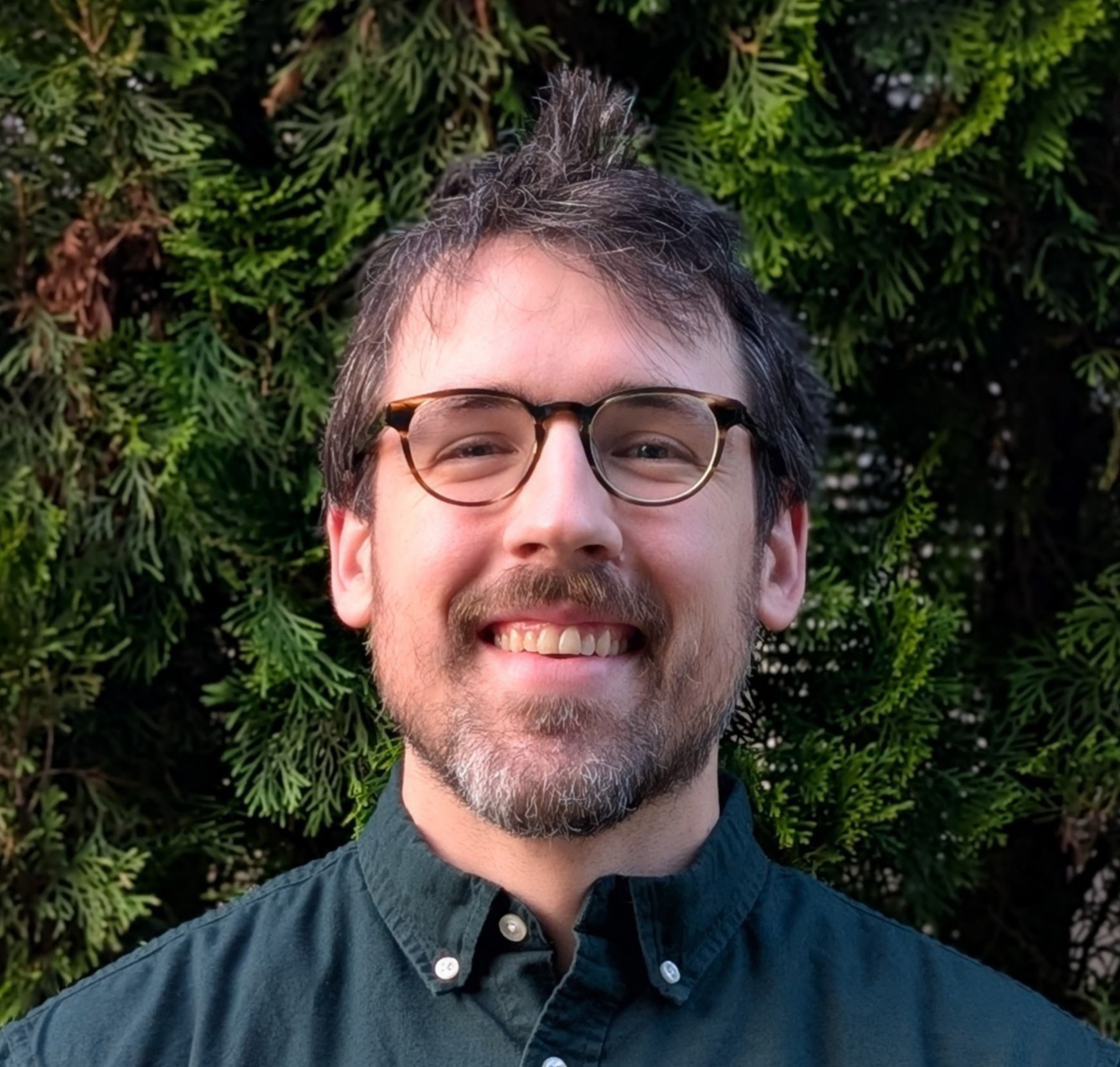 An image of Clay Whetung. He is wearing a green shirt and glasses and is standing in from of a tree